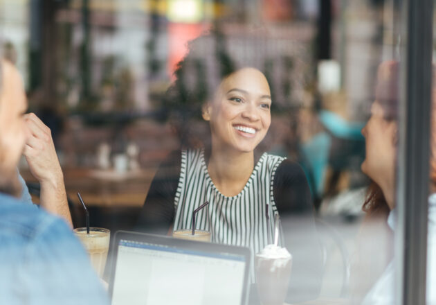 A woman visible through a glass window laughs with a group of friends at a table.