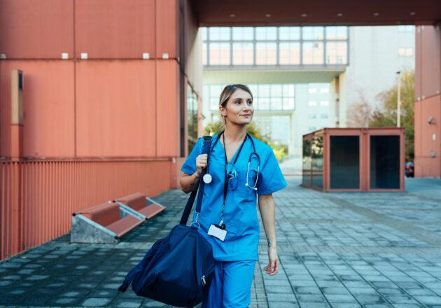 A nurse in blue scrubs walks down a pathway while holding a duffle bag on her shoulder.