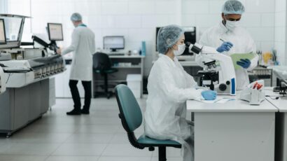In a lab, one scientist peers into a microscope and another writes on a clipboard.